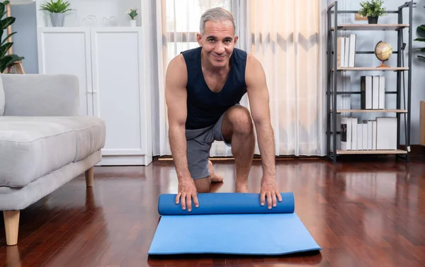 Active and sporty senior man preparing, rolling fitness exercising mat on living room floor at home. Home exercise as concept of healthy fit body lifestyle after retirement. Clout