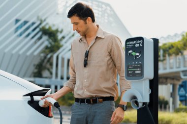 Genç adam, şehrin ticari otoparkındaki şarj istasyonundan elektrikli araba aküsünü şarj etmek için EV şarj aleti koymuş. Sürdürülebilir çevre dostu şehir yolculuğu için şarj edilebilir EV araba. Yararlı