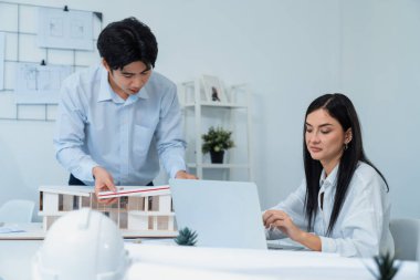 Profesyonel Asyalı erkek mimar ev modelinin uzunluğunu ölçmek için cetvel kullanırken, beyaz tenli genç meslektaşı ev modeliyle toplantı masasındaki verileri analiz etmek için dizüstü bilgisayar kullanıyor. Tertemiz..
