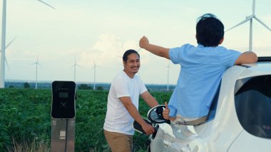 İlerici mutlu aile kavramı elektrikli araçla rüzgar çiftliğinde eğleniyor. Rüzgar türbini jeneratöründen gelen temiz yenilenebilir enerji ile şarj edilen elektrikli araç..