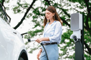 Yolculuk sırasında doğal orman ya da milli parktaki elektrikli araba için şarj olan genç bir kadın. Tatil ve tatil boyunca çevre dostu seyahat. Yükselt