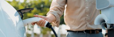 Genç adam şehrin ticari otoparkındaki şarj istasyonundan elektrikli araba aküsünü şarj ediyor. Sürdürülebilir çevre dostu şehir yolculuğu için şarj edilebilir EV araba. Panorama Çıkarı