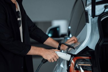 Genç adam EV elektrikli arabasıyla alışveriş merkezinin otoparkına gidiyor şehir merkezindeki şarj oluyor yeşil, temiz, yeniden şarj edilebilir elektrikli araç iç organları ile kentsel sürdürülebilirlik yaşam tarzını gösteriyor