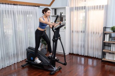 Enerjik ve güçlü atletik Asyalı kadın evde eliptik koşu makinesiyle koşuyor. Formda fiziğin takibi ve ev çalışması ve eğitimle sağlıklı yaşam tarzına bağlılık. Kuvvetli