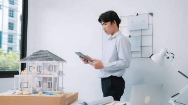Profesyonel yakışıklı Asyalı proje yöneticisi ev modelini kontrol etmek için tablet kullanırken ev modelinin yapısını dikkatlice inceleyip arkasında planlar asılıydı. Yaratıcı tasarım. Tertemiz..