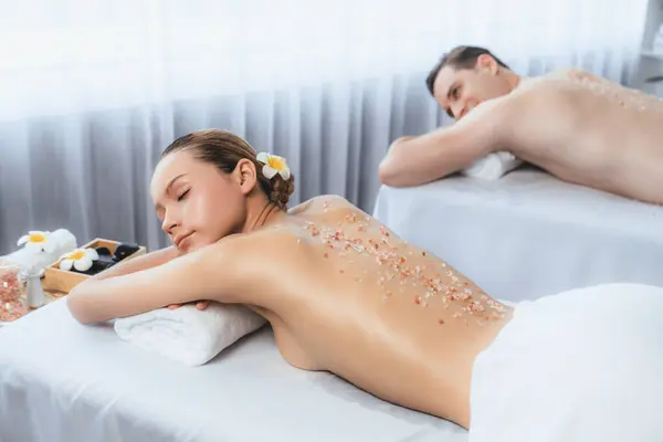 Blissful couple customer having exfoliation treatment in luxury spa salon with warmth candle light ambient. Salt scrub beauty treatment in health spa body scrub. Quiescent