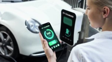 Akıllı telefon akü durumu arayüzünü elinde tutan iş kadını akıllı EV mobil uygulama ile otomobil parkındaki şarj istasyonundan elektrik şarj eden çevre dostu EV araba. İnceleyin