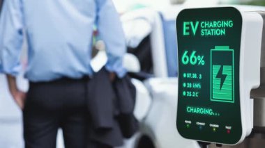 Şehir merkezindeki şarj istasyonundan ya da bulanık arka plandaki kamu parkından elektrikli araba şarj ettikten sonra bekleyen bir işadamı. Çevre dostu şarj edilebilir EV araba alternatif temiz enerji kullanır. İnceleyin