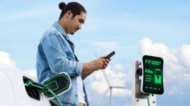 Asyalı bir adam rüzgar türbini çiftliğindeki şarj istasyonundan elektrik şarj ederken akıllı telefon kullanıyor. Alternatif sürdürülebilir ve temiz enerji kullanımı kavramı. İnceleyin