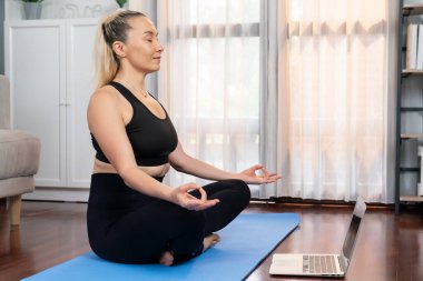 Spor giyim bölümündeki son sınıf kadın evde egzersiz paspası üzerinde meditasyon pozisyonunda yoga yapıyor. Huzurlu ve huzurlu bir huzur içinde sağlıklı bir emekli yaşam tarzı. Etki