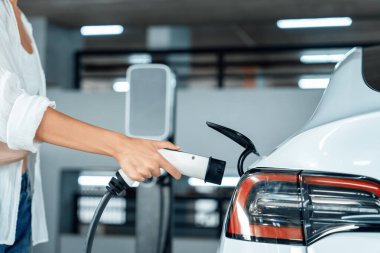 Genç bir kadın EV elektrikli arabasıyla alışveriş merkezinin otoparkına gidiyor şehir merkezinde elektrik iç organlarının yeşil temiz şarj edilebilir enerji ile sürdürülebilirlik yaşam tarzını gösteriyor.