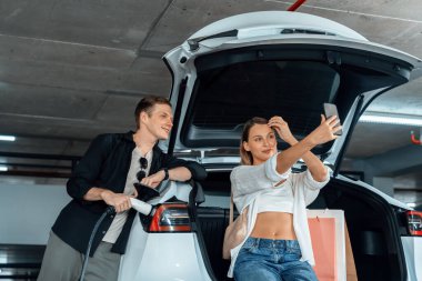 Genç çift EV elektrikli arabasıyla alışveriş merkezinin otoparkına gidiyor şehir merkezindeki şarj oluyor yeşil temiz şarj edilebilir elektrikli araç iç organları ile kentsel sürdürülebilirlik yaşam tarzını gösteriyor