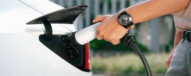 Genç adam şehrin ticari otoparkındaki şarj istasyonundan elektrikli araba aküsünü şarj ediyor. Sürdürülebilir çevre dostu şehir yolculuğu için şarj edilebilir EV araba. Panorama Çıkarı