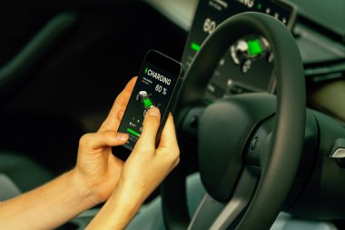 Aracın içindeki genç kadın, modern EV arabasındaki akıllı telefon ekranında akü şarj etme uygulamasını kontrol ediyor. Yükselt