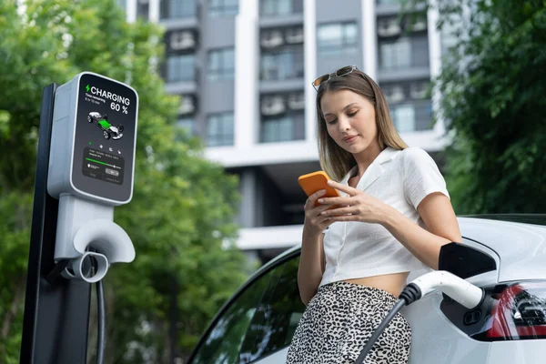 Young woman travel with EV electric car charging in green sustainable city outdoor garden in summer. Urban sustainability lifestyle by green clean rechargeable energy of electric BEV vehicle innards