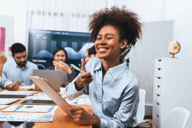 Bir grup ofis çalışanı ile birlikte arka planda ekranda gösterim paneliyle buluşan mutlu Afrikalı genç bir iş kadını portresi. Takım toplantısında kendine güveni tam bir ofis hanımı. Uyum