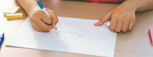 A cropped image of female businesswoman hand writing marketing on paper with pencil and sticky notes placed on table. Creative start up business and teamwork concept. Focus on hand. Variegated.