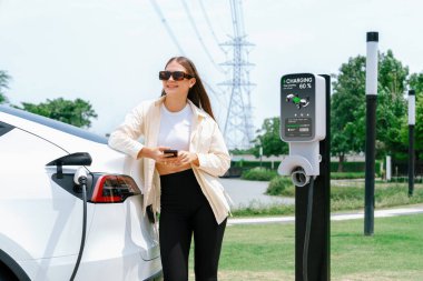 Genç bir kadın EV otomobil şarj istasyonu Green City Park 'ta elektrik ödemek için akıllı telefonu kullanıyor. Modern çevre ve EV araçlı sürdürülebilir şehir yaşam tarzı. Yararlı