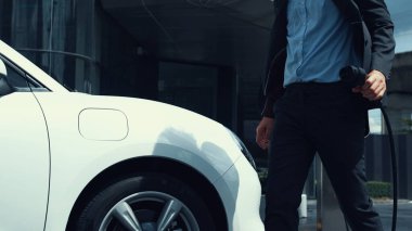 İş adamı elektrikli arabasını şehir merkezindeki şarj istasyonundan ya da otoparktan şarj ediyor. Şehir yaşam tarzında alternatif temiz enerji kullanan çevre dostu şarj edilebilir araba. İnceleyin.