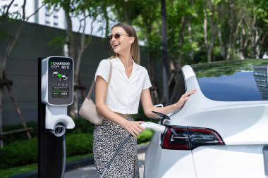 Genç bir kadın yazın yeşil sürdürülebilir şehir bahçesinde EV elektrikli arabasıyla seyahat ediyor. Kentsel sürdürülebilirlik yaşam tarzı yeşil temiz yeniden şarj edilebilir enerji elektrikli BEV araç iç organları