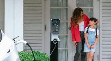 Küçük mutlu kız, annesinin EV şarj istasyonundan elektrikli aracı şarj etmesine yardım ederken çevre dostu ve enerji sürdürülebilirliğini öğreniyor. EV araba ve modern aile. Panorama Eşzamanlaması