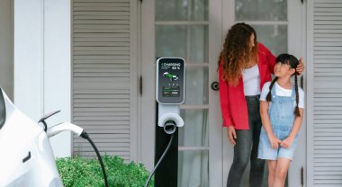 Happy little young girl learn about eco-friendly and energy sustainability as she help her mother recharge electric vehicle from home EV charging station. EV car and modern family. Panorama Synchronos clipart