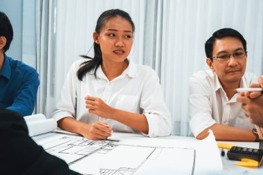 Mimari proje üzerinde birlikte çalışan çeşitli inşaat mühendisi ve müvekkil grupları, inşaat planını gözden geçirip toplantı masasında plan oluşturuyorlar. Tedbirli