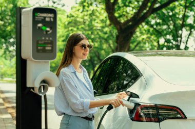 Yolculuk sırasında doğal orman ya da milli parktaki elektrikli araba için şarj olan genç bir kadın. Tatil ve tatil boyunca çevre dostu seyahat. Yükselt