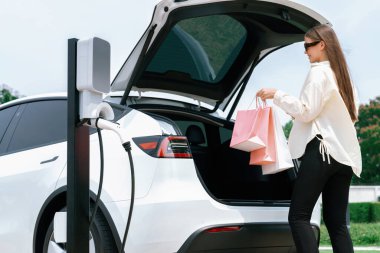 Elinde alışveriş çantası olan genç bir kadın Green City Park 'taki şarj istasyonundan EV araba aküsünü şarj ediyor. Modern kadın çevre dostu elektrikli araç ile alışveriş yapıyor. Yararlı