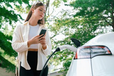 Genç bir kadın EV otomobil şarj istasyonu Green City Park 'ta elektrik ödemek için akıllı telefonu kullanıyor. Modern çevre ve EV araçlı sürdürülebilir şehir yaşam tarzı. Yararlı