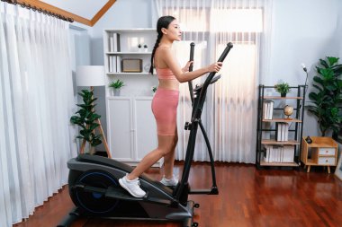 Enerjik ve güçlü atletik Asyalı kadın evde eliptik koşu makinesiyle koşuyor. Formda fiziğin takibi ve ev çalışması ve eğitimle sağlıklı yaşam tarzına bağlılık. Kuvvetli
