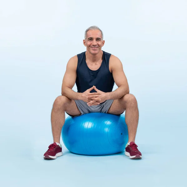 Full body length shot athletic and sporty senior man with fitness exercising ball in sitting posture on isolated background. Healthy active and body care lifestyle after retirement. Clout