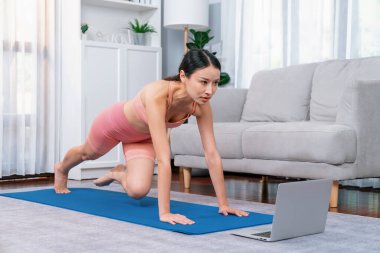 Spor kıyafetli Asyalı bir kadın antrenman rutini olarak spor minderinin üzerinde burpee yapıyor. Çekici kız sağlıklı yaşam tarzını online egzersiz videosuyla takip ediyor. Kuvvetli