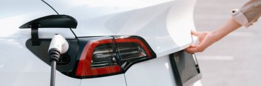 Otoparktaki şarj istasyonunun pilini şarj ederken genç bir kadın bagajı açıyor. Sürdürülebilir çevre dostu seyahat için alternatif enerji ve şarj edilebilir elektrikli araç.
