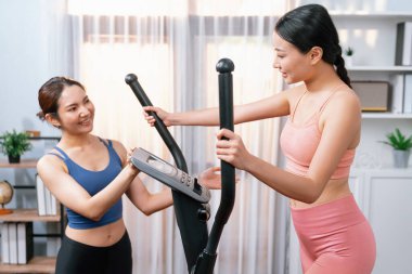 Enerjik ve güçlü atletik Asyalı kadın evde spor arkadaşı ya da antrenörü ile birlikte eliptik koşu makinesiyle koşuyor. Formda fiziğin takibi ve sağlıklı yaşam tarzına bağlılık. Kuvvetli