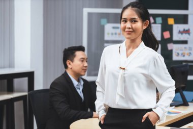 Modern ofiste gülümserken kendine güvenen beyaz üniformalı başarılı bir iş kadını. Kollarında akimbo olan Asyalı kadın portresi. Ortağı bilgisayar entelektüeli üzerinde çalışıyor..