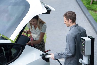 Genç çift yazın yeşil sürdürülebilir şehir bahçesinde EV elektrikli araba ile seyahat ederken, yeşil temiz yeniden şarj edilebilir elektrikli araç iç organları ile kentsel sürdürülebilirlik yaşam tarzını gösteriyor.