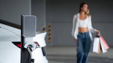Genç bir kadın EV elektrikli arabasıyla alışveriş merkezinin otoparkına gidiyor şehir merkezinde elektrik iç organlarının yeşil temiz şarj edilebilir enerji ile sürdürülebilirlik yaşam tarzını gösteriyor.