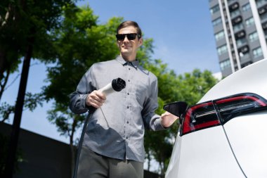 Genç adam yazın yeşil sürdürülebilir şehir bahçesinde EV elektrikli araba ile seyahat ederek yeşil, temiz, yeniden şarj edilebilir elektrikli araç iç organları ile kentsel sürdürülebilirlik yaşam tarzını gösteriyor