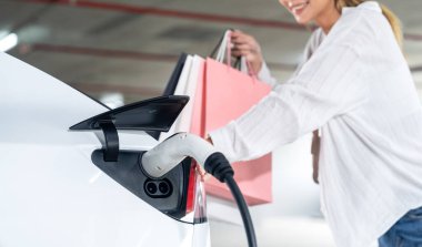 Genç bir kadın EV elektrikli arabasıyla alışveriş merkezinin otoparkına gidiyor şehir merkezinde elektrik iç organlarının yeşil temiz şarj edilebilir enerji ile sürdürülebilirlik yaşam tarzını gösteriyor.