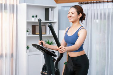 Enerjik ve güçlü atletik Asyalı kadın evde eliptik koşu makinesiyle koşuyor. Formda fiziğin takibi ve ev çalışması ve eğitimle sağlıklı yaşam tarzına bağlılık. Kuvvetli