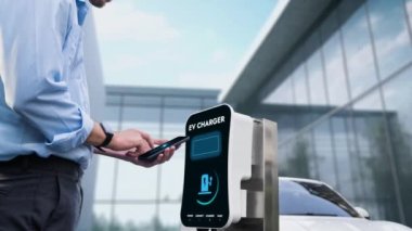 Modern iş adamı akıllı telefonun 3D hologramından batarya durumunu kontrol ederken EV 'si şarj olan enerjisini kontrol ediyor. Gelecekteki EV otomotiv teknolojisinin yenilikçi teknolojik gelişimi. İnceleyin