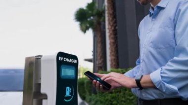 İşadamı, EV batarya durum hologramını akıllı telefon uygulamasından kontrol ederken EV aracını EV 'nin şarj ettiği çevre yeşil şehir parkından ağaçla şarj ediyor. Geleceğin yenilikçi enerji sürdürülebilirliği. İnceleyin