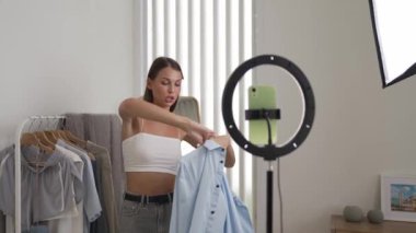 Genç sosyal medyum kadın moda videosu yapıyor. Blogcu kameraya selfie çubuğu ve ışık halkası kullanarak gülümserken, aynı zamanda ikna edici çevrimiçi kıyafetlerin izleyicilere veya takipçilere vlog satmasını sağlıyor. Düzenle
