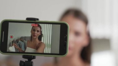 Genç sosyal medyum kadın moda videosu yapıyor. Blogcu kameraya selfie çubuğu ve ışık halkası kullanarak gülümserken, aynı zamanda ikna edici çevrimiçi kıyafetlerin izleyicilere veya takipçilere vlog satmasını sağlıyor. Düzenle