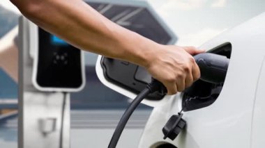 Adam elektrikli araca akıllı EV şarj cihazı takıyor ve EV şarj istasyonundan modern tasarım binası ya da arka planda evi olan şarj oluyor. Geleceksel sürdürülebilir güç ve EV araba kullanımı. İnceleyin