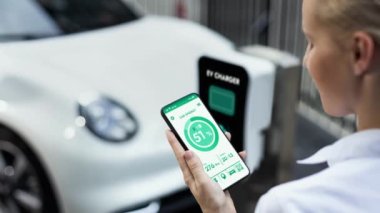 Akıllı telefon akü durumu arayüzünü elinde tutan iş kadını akıllı EV mobil uygulama ile otomobil parkındaki şarj istasyonundan elektrik şarj eden çevre dostu EV araba. İnceleyin
