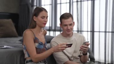 Genç çift, kredi kartıyla ödemek için internet üzerinden ödeme uygulaması ve dijital cüzdan kullanarak yatak odasında oturuyor. Mobil internet üzerinden alışveriş ve modern alımlar. Düzenle