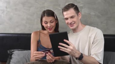Genç çift, kredi kartıyla ödeme yapabilmek için internet üzerinden ödeme uygulaması ve dijital cüzdan kullanarak yatak odasında oturuyor. Mobil internet üzerinden alışveriş ve modern alımlar. Düzenle
