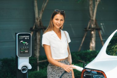 Genç bir kadın yazın yeşil sürdürülebilir şehir bahçesinde EV elektrikli arabasıyla seyahat ediyor. Kentsel sürdürülebilirlik yaşam tarzı yeşil temiz yeniden şarj edilebilir enerji elektrikli BEV araç iç organları
